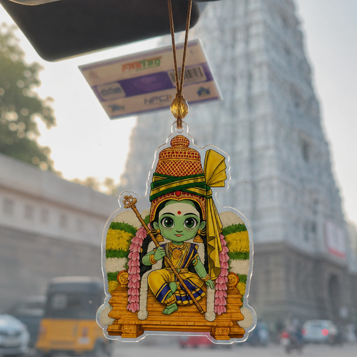 A colorful acrylic hanging ornament featuring a cartoon depiction of Lord Hanuman, suspended by a cord from a car windshield with a blurred street scene in the background.